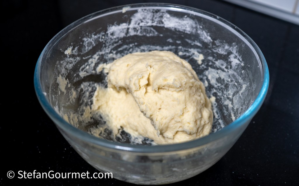 A glass bowl containing a mound of dough with a slightly sticky texture, placed on a dark countertop.