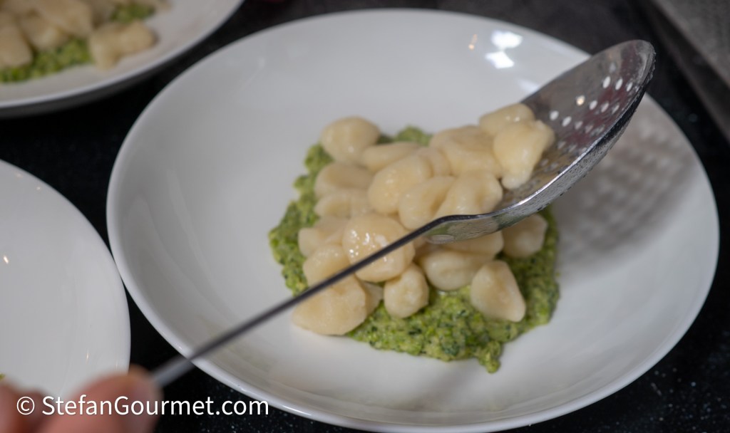 A spoon is serving soft gnocchi onto a bed of vibrant green pesto on a white plate.