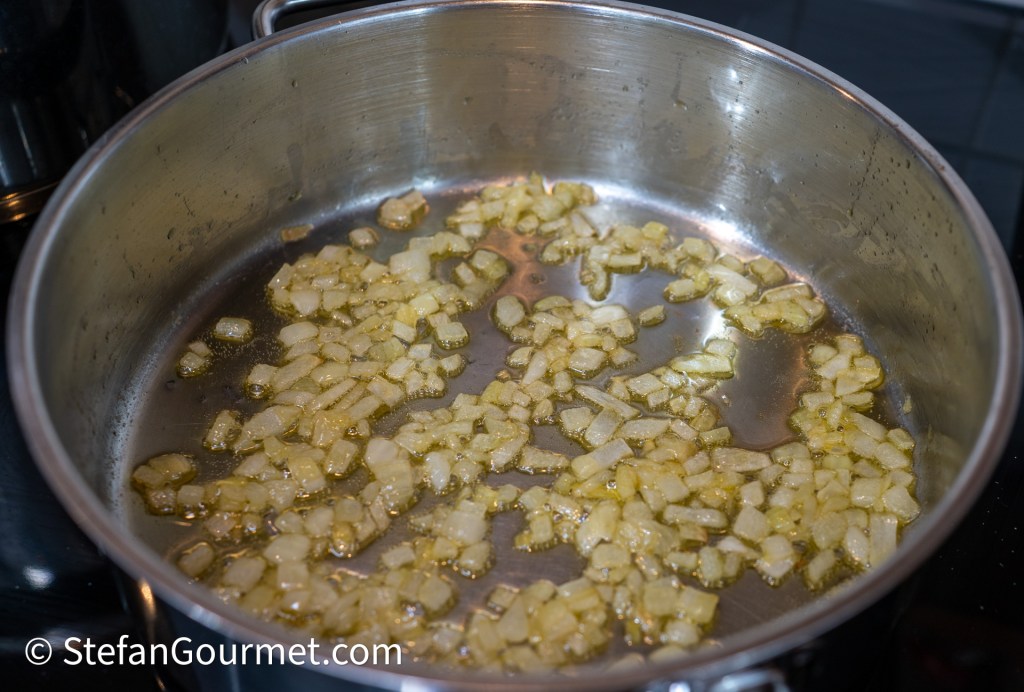 Diced onions sautéing in a silver pot with oil on a stovetop.