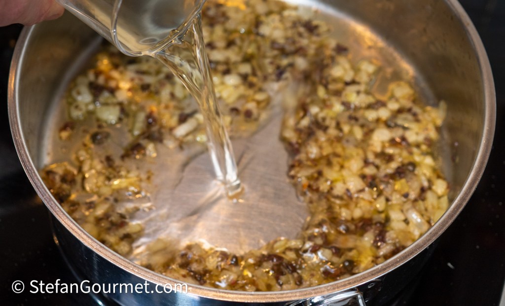 A hand pouring liquid into a pan containing sautéed onions and herbs on a stove.