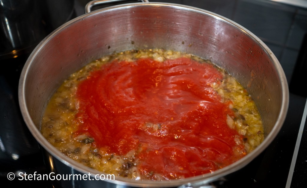 A pot on a stovetop containing sautéed onions and garlic topped with a layer of red tomato sauce.