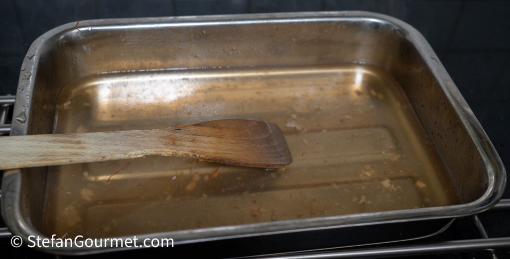 A stainless steel roasting pan with some liquid and residue at the bottom, accompanied by a wooden spoon resting inside.