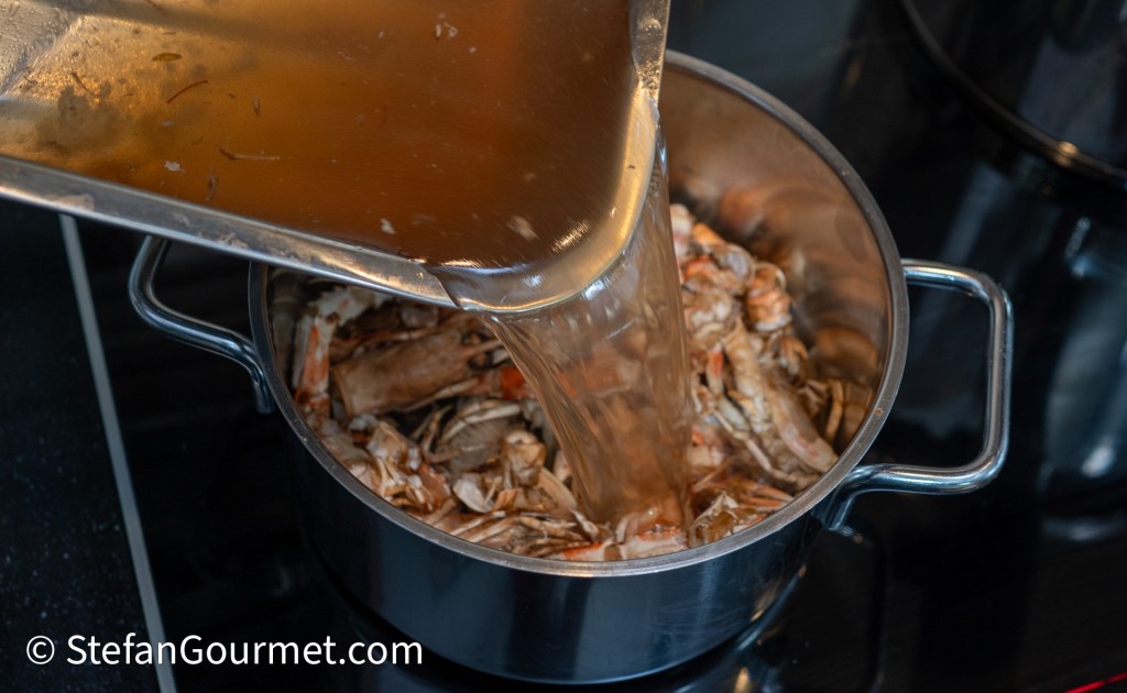 A pot filled with cooked seafood, receiving broth being poured in from a metal container.