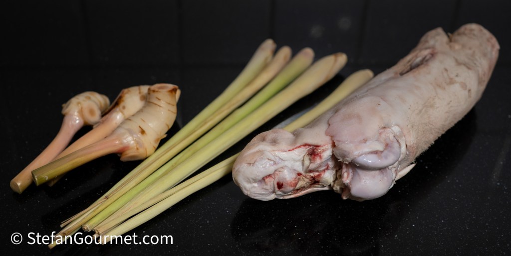 Raw lemongrass stalks and galangal root beside a cut piece of pork belly on a dark surface.