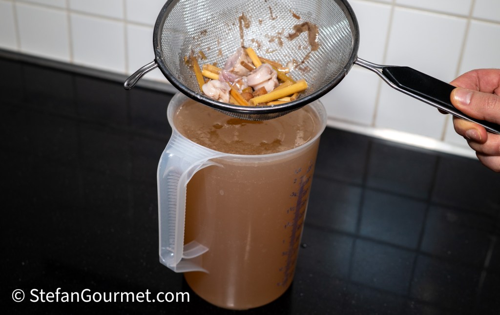 A person using a fine mesh strainer to filter solid ingredients from broth into a measuring pitcher on a kitchen counter.