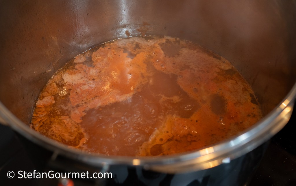 A close-up view of simmering broth in a metal pot, showing a rich, brownish liquid with flecks of seasoning and bubbles on the surface.