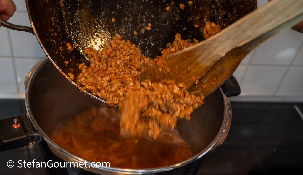 A wooden spoon scoops out cooked food, pouring it into a pot on the stove. The food appears to be a mixture of grains and spices.