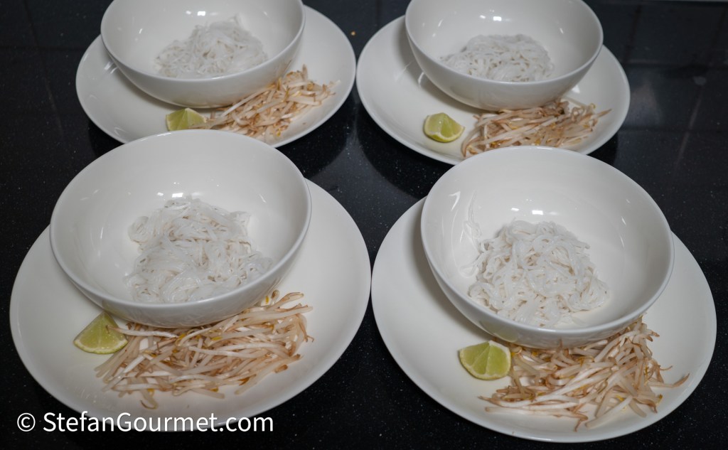 Four white bowls with rice noodles served on white plates, accompanied by bean sprouts and lime wedges.