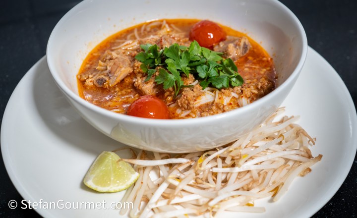 A bowl of spicy noodle soup with tender meat, cherry tomatoes, and fresh cilantro, served with a side of bean sprouts and a lime wedge.