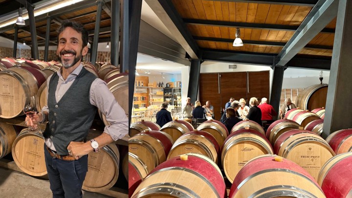 A smiling man holding a glass of wine stands among wooden wine barrels in a winery.