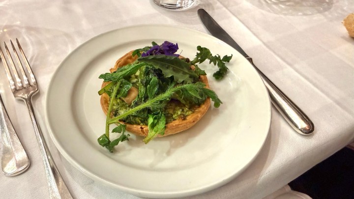 A small tart filled with green pesto and topped with fresh greens and an edible flower, placed on a white plate with cutlery beside it.
