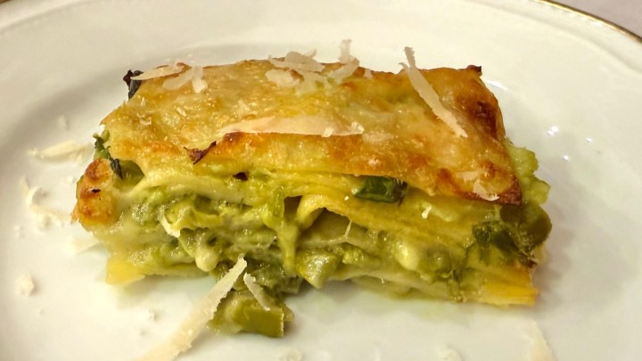A slice of creamy green vegetable lasagna topped with melted cheese shavings on a white plate.