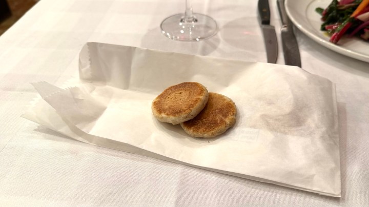 Two small, round, toasted bread muffins on a white paper napkin, set on a table with a wine glass and a plate of colorful vegetables in the background.
