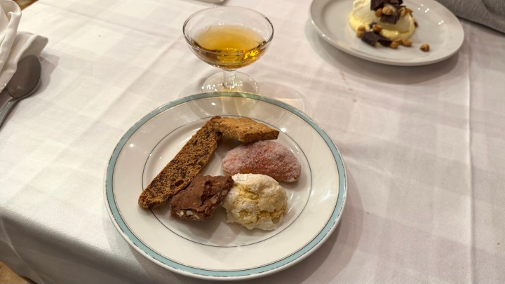 A plate with various types of cookies, including a chocolate cookie, a nutty cookie, and a powder-coated cookie, accompanied by a small glass of amber-colored drink.