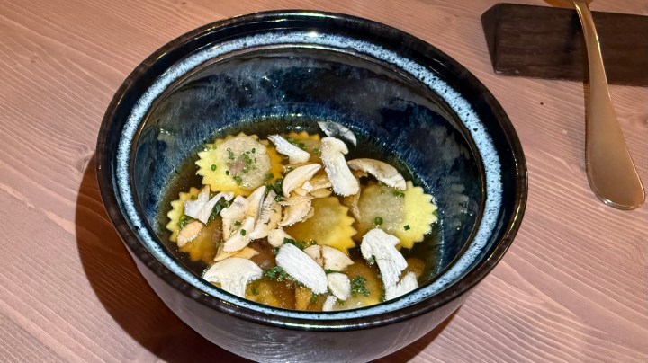 A bowl of ravioli in clear broth topped with sliced mushrooms and herbs.