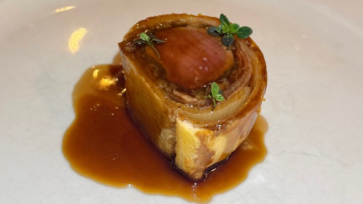 A cylindrical piece of cooked meat garnished with small green herbs, served on a white plate with a rich brown sauce.