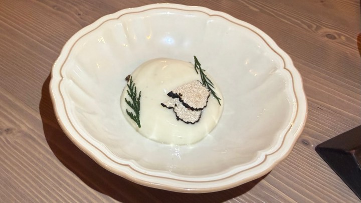 A creamy dessert presented in a shallow white bowl, garnished with black truffle slices and green herbs.
