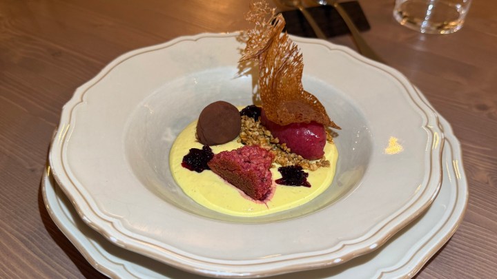 A gourmet dessert featuring a vibrant arrangement of various components, including a purple sorbet, a chocolate truffle, a berry compote, pieces of cake, and a decorative caramel lace, all served on a creamy yellow base in a white dish.