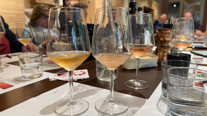 A table setting showing several wine glasses with light-colored liquid, surrounded by other glasses and dining items. In the background, blurred figures of people engaged in conversation can be seen.