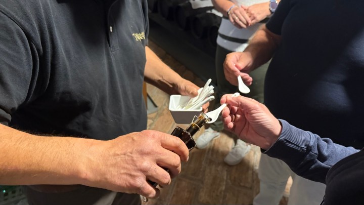 A person pouring a dark liquid from a bottle into a small dish, while another person holds a spoon. Several hands are visible, indicating a tasting or sampling event.