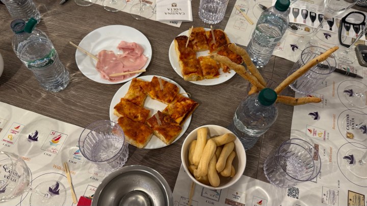 A table setting featuring various food items, including slices of pizza, ham, breadsticks, and a bowl of snacks, accompanied by several bottles of water and glassware.