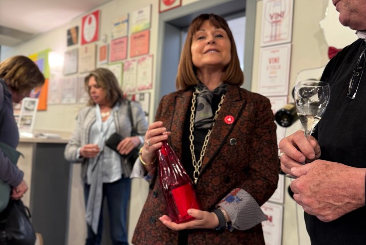 A woman holding a bottle of red wine, speaking to a group of people in a room with wall certificates and awards visible in the background.