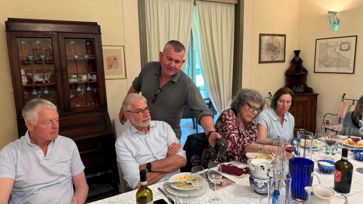A group of five people seated at a dining table, enjoying a meal. One person is pouring wine, while others are engaged in conversation. The setting includes a light-colored room with a wooden cabinet and decorative items in the background.