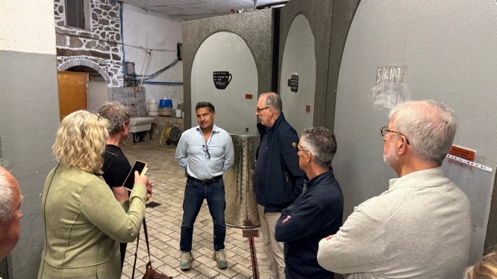 A group of people gathered in a winery, engaged in a discussion with a speaker in front of large wine fermentation tanks.