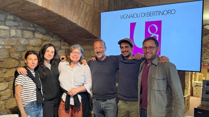Group of six people posing together in a rustic space with stone walls, smiling at the camera. A screen in the background displays the text 'Vignaioli di Bertinoro'.