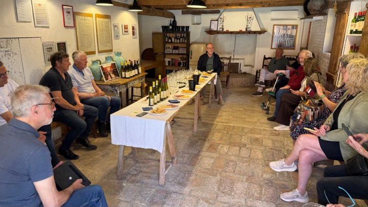 A group of people seated around a long table in a cozy tasting room, with wine bottles and food laid out for a tasting event.