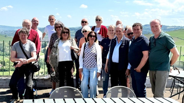 A group of 18 people standing together outdoors with a scenic landscape in the background, featuring rolling hills and blue skies.