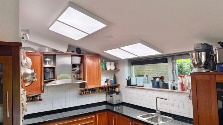 A modern kitchen featuring wooden cabinets, a sleek black countertop, and large overhead lights. The room includes various kitchen appliances, glassware, and organized spice jars, with a window providing natural light.