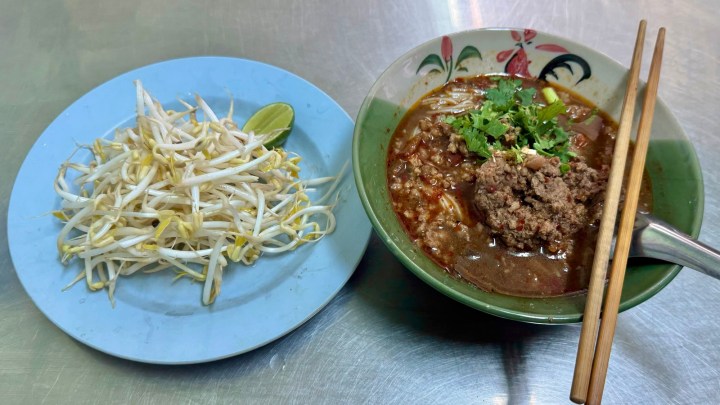 A bowl of stew with meat, topped with chopped cilantro, served alongside a plate of fresh bean sprouts and a lime wedge.