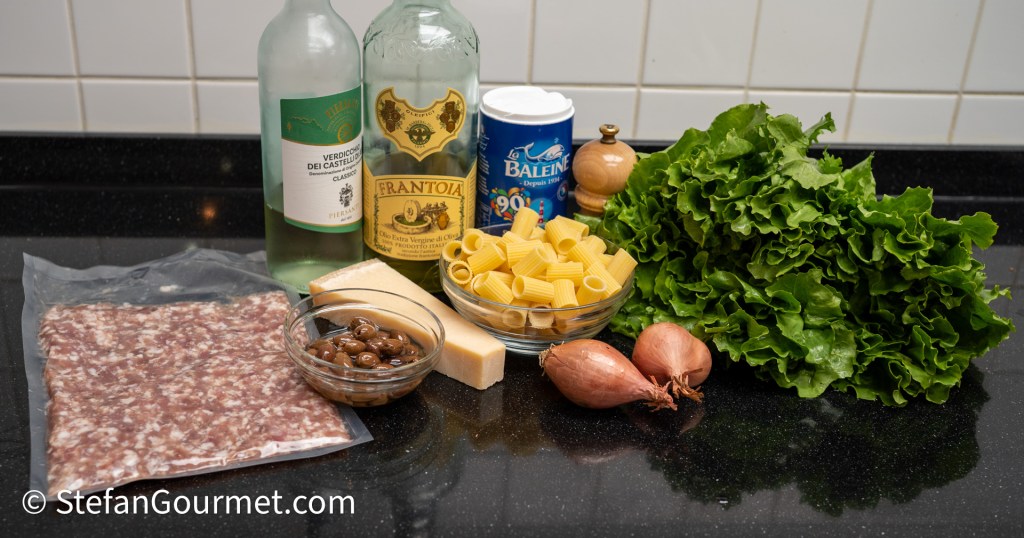 An assortment of ingredients for cooking displayed on a black countertop, including a package of ground meat, two bottles of wine, a bowl of pasta, a bowl of olives, a block of cheese, a container of salt, fresh lettuce, and two shallots.