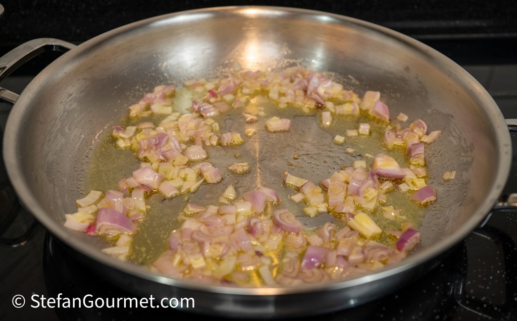 Chopped shallots sautéing in olive oil in a stainless steel skillet on a stovetop.