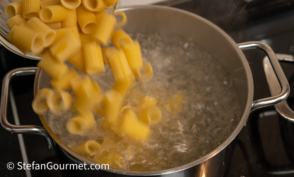 A pot of boiling water with uncooked rigatoni pasta being poured in from a spoon.