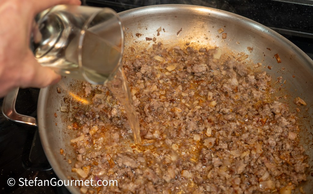 A hand pouring liquid from a glass into a pan with cooked ground meat and onions on a stovetop.
