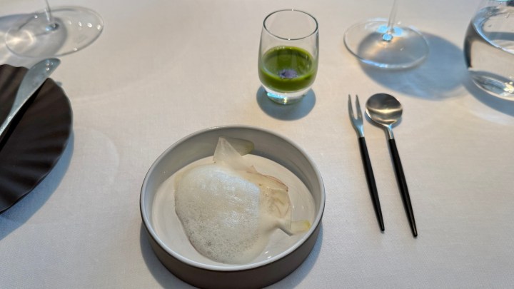 A plated dish featuring a delicate foam on top, garnished with translucent elements, accompanied by a small glass of green liquid and silver cutlery on a white tablecloth.
