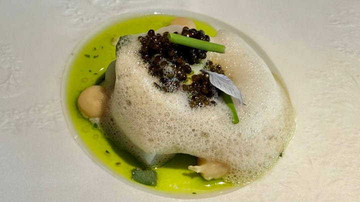 A gourmet dish featuring a white protein, possibly fish, topped with caviar and herbs, served in a vibrant green sauce, with a foamy garnish and small garnishes of vegetables.