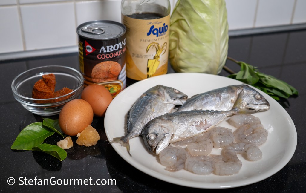 A selection of cooking ingredients including three whole fish, shrimp, eggs, coconut milk, a bottle of fish sauce, a head of cabbage, curry paste, and curry leaves arranged on a dark countertop.
