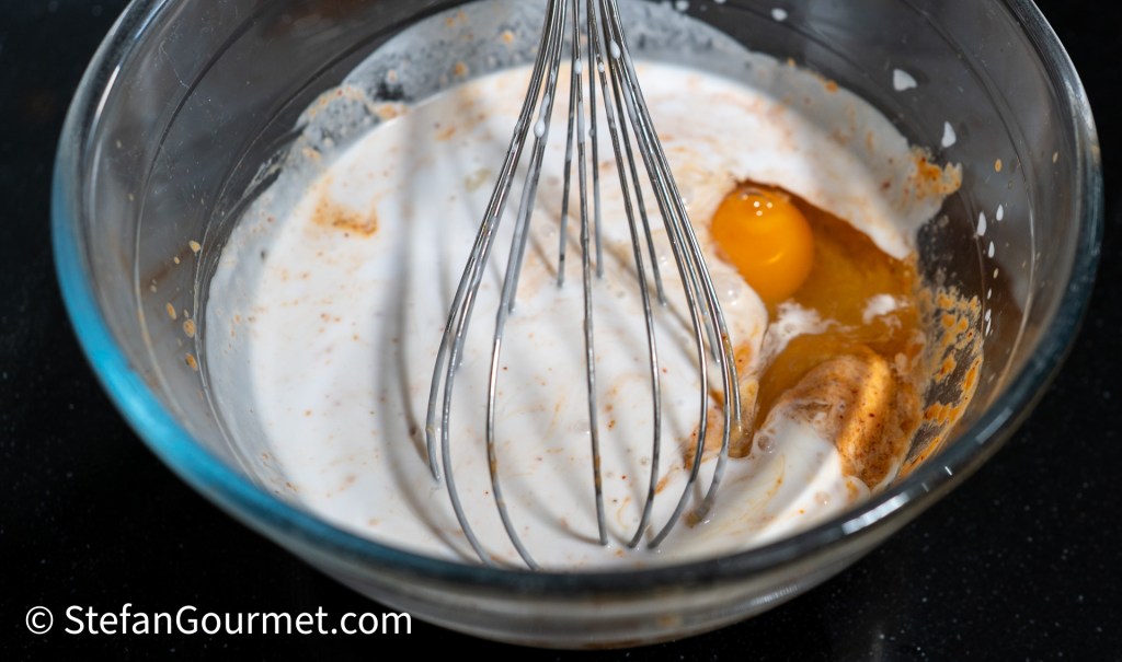 A glass bowl containing a mixture of cream, an egg, and some spices, with a whisk resting inside.