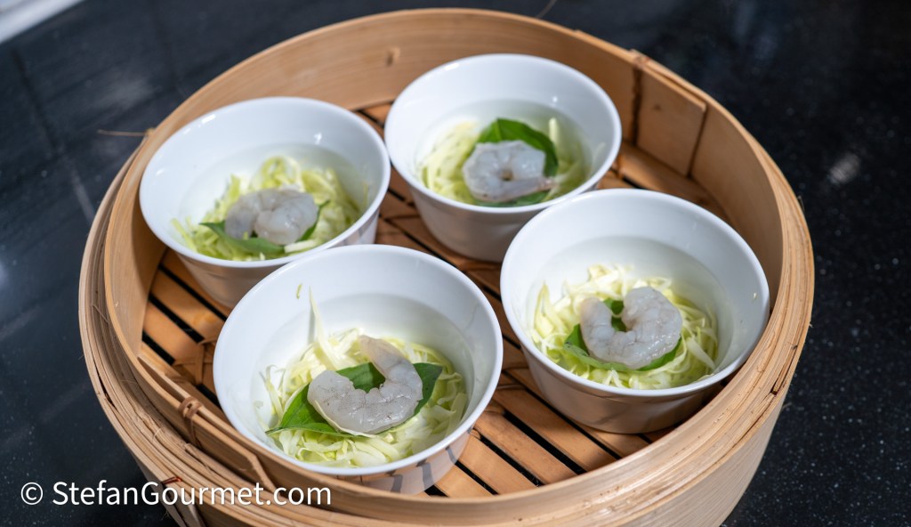 Four small white bowls containing shrimp, placed on a bed of cabbage and garnished with green leaves, arranged in a bamboo steamer.