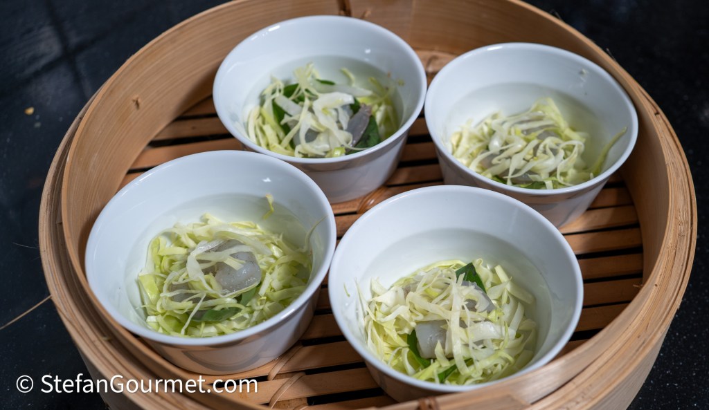 Steamer basket containing four small white bowls filled with cabbage, herbs, and pieces of seafood in a light broth.