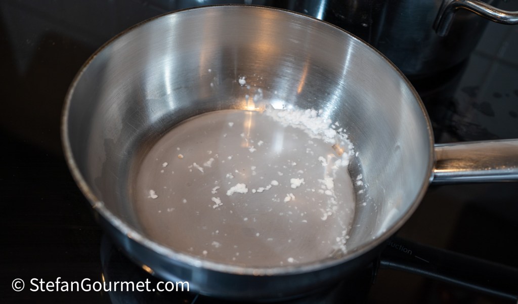 A close-up of a stainless steel saucepan with a small amount of water and residue at the bottom, placed on a black induction cooktop.