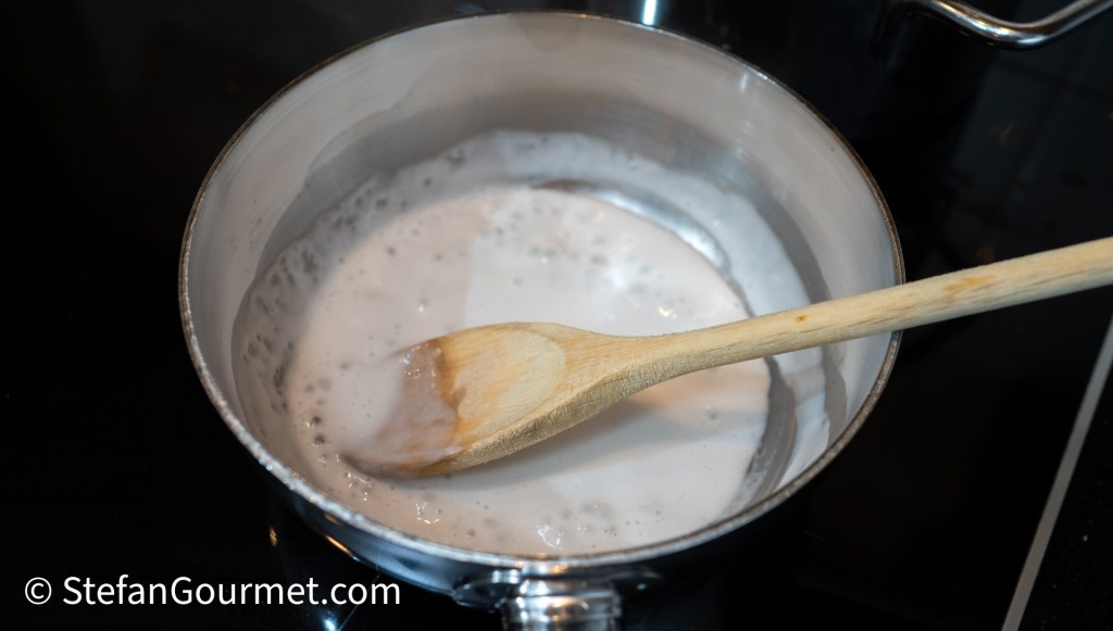 A saucepan filled with a creamy liquid being stirred with a wooden spoon, on a black stovetop.