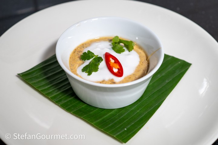A small white bowl filled with creamy soup, topped with coconut cream, fresh cilantro, and a slice of red chili, resting on a green banana leaf on a white plate.