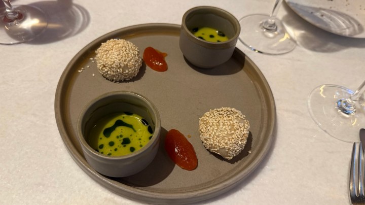 A round plate featuring two small bowls of green sauce and three sesame-coated balls alongside red sauce drizzles.