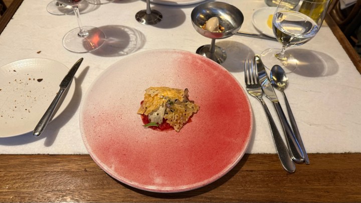 A elegantly plated dish on a large, red-accented plate, featuring a small serving of food topped with a golden, crispy garnish, surrounded by dining utensils and glasses of wine and water.