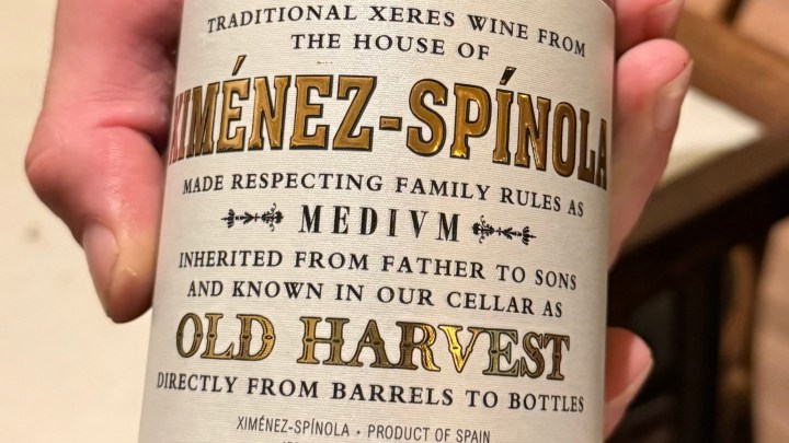 Close-up of a hand holding a wine bottle label from Ximénez-Spínola, featuring details about traditional xeres wine, including the words 'MEDIVM' and 'OLD HARVEST'.