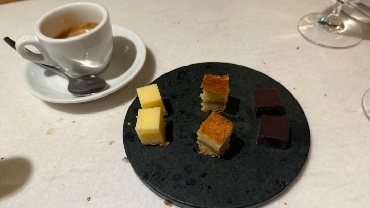 A plate featuring four small dessert squares: two yellow, one golden, and two dark chocolate squares, alongside a cup of espresso.